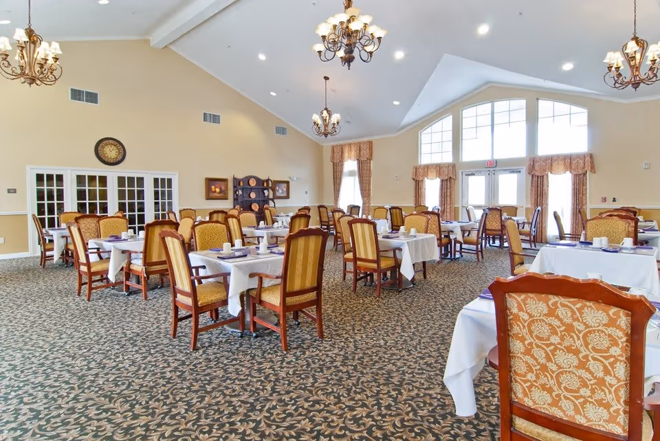 A spacious dining room with multiple tables covered in white tablecloths, each set with cups and napkins. The room features patterned carpet, high vaulted ceilings with chandeliers, large windows with curtains, and a clock on the wall above double doors.