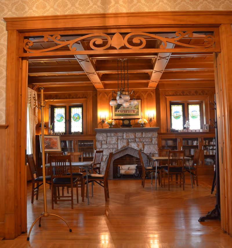 A cozy dining room with wooden floors and walls, featuring a stone fireplace in the center. The room has several wooden tables and chairs, stained glass windows on either side of the fireplace, and a vintage chandelier hanging from the ceiling. Decorative wooden trim and built-in shelves with dishes are visible.