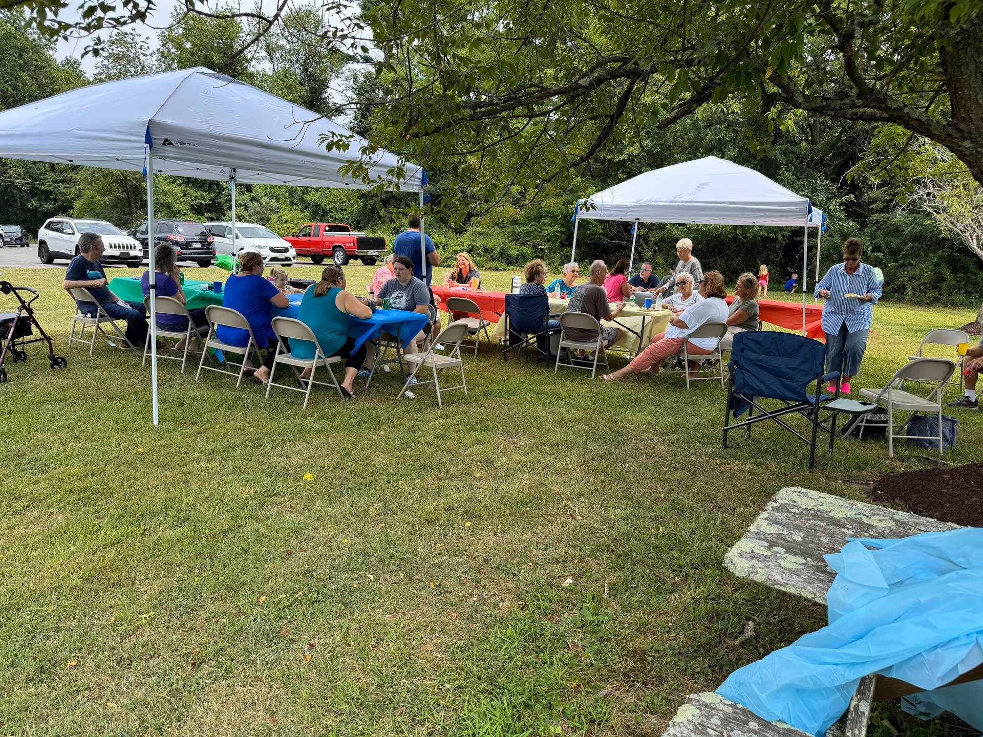 A group of people sitting and socializing under two white canopy tents set up on a grassy area with trees in the background. Several folding chairs and tables with colorful tablecloths are arranged under the tents. Cars are parked in the background, and a few people are standing or walking nearby.