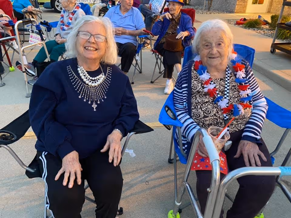 Two elderly women sitting outdoors on folding chairs, smiling at the camera. One woman wears a dark blue sweater with a decorative necklace, and the other wears a striped cardigan with a red, white, and blue lei around her neck, holding a drink. Other elderly people are seated in the background, some with walkers, enjoying a sunny day at what appears to be a social gathering.