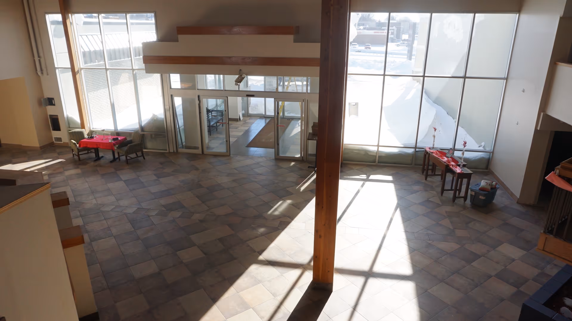 Spacious lobby area with large windows letting in natural light, tiled floor, a wooden pillar in the center, a small table with two chairs on the left, and a table with decorative items on the right near the windows.
