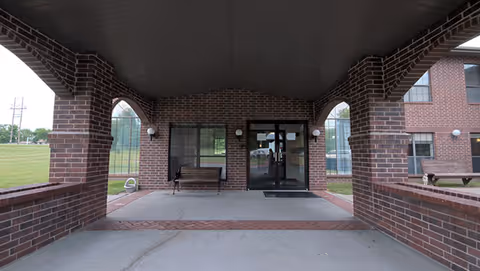 Covered entrance area of a brick building with arched openings on the sides, two benches, and glass double doors leading inside. The building is surrounded by grass and has windows visible on the right side.