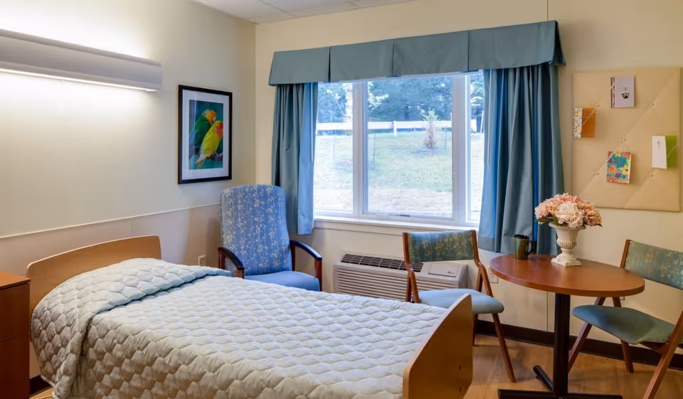 A well-lit bedroom in a senior living facility featuring a single bed with a light blue quilted bedspread, a blue upholstered armchair, two wooden chairs with blue cushions, a round wooden table with a vase of pink flowers, a large window with blue curtains, a framed picture of colorful birds on the wall, and a beige bulletin board with pinned cards.
