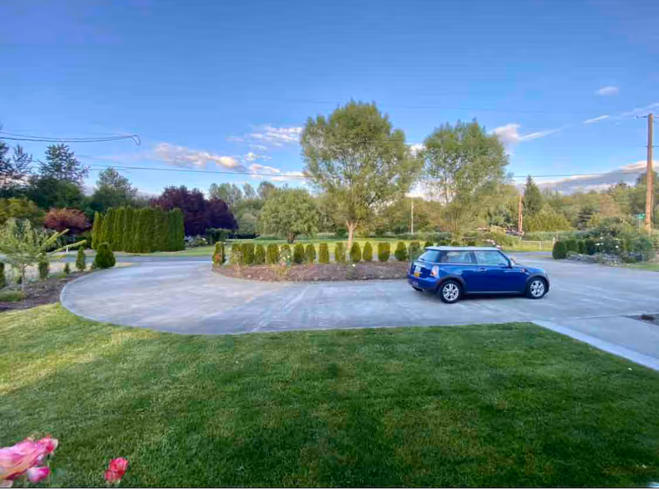 A circular driveway with a blue Mini Cooper parked on it, surrounded by green grass, trees, and shrubs under a clear blue sky.