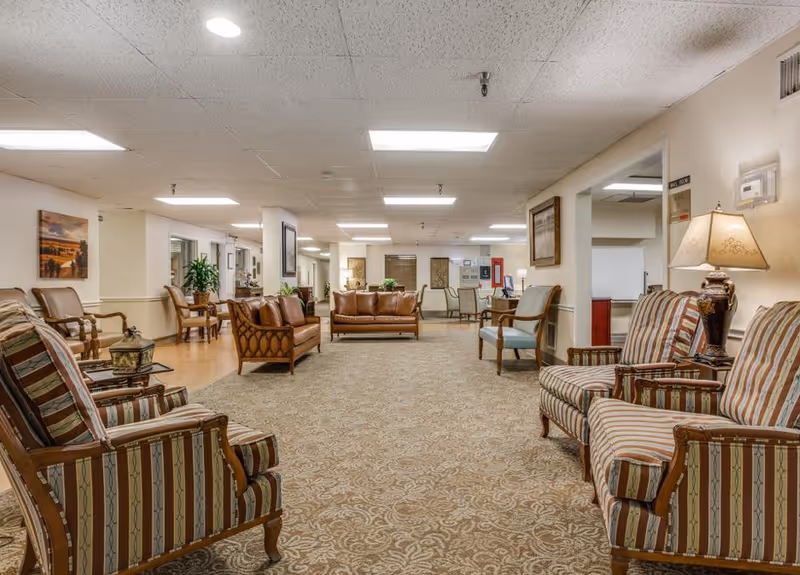 A spacious senior living facility common area with patterned carpet and multiple seating arrangements including striped armchairs, leather sofas, and wooden chairs. The room is well-lit with ceiling lights and decorated with framed artwork and a table lamp.