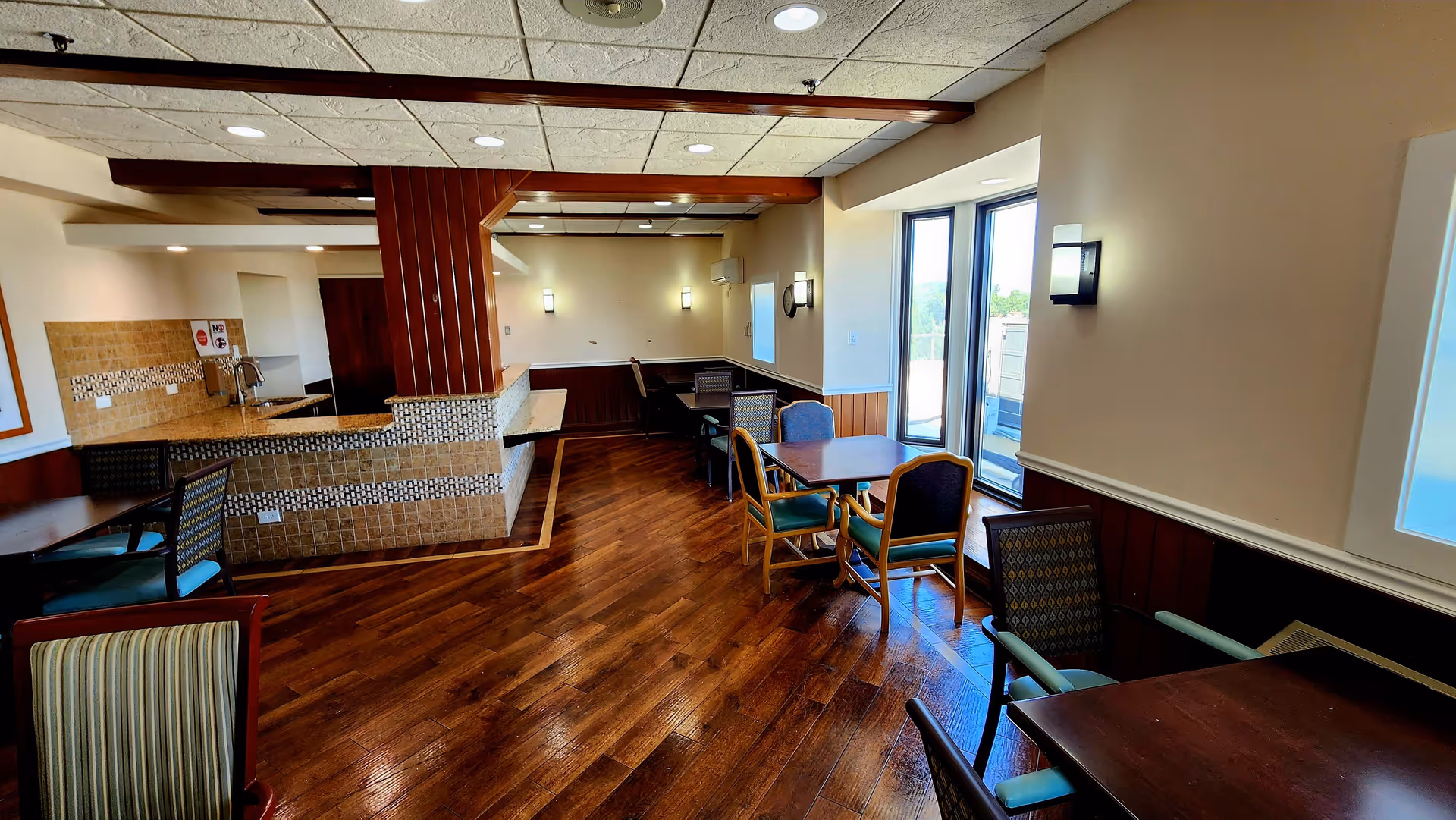 Interior view of a dining area in a senior living facility with wooden floors, multiple tables and chairs, a counter with a tiled front, and large windows letting in natural light.