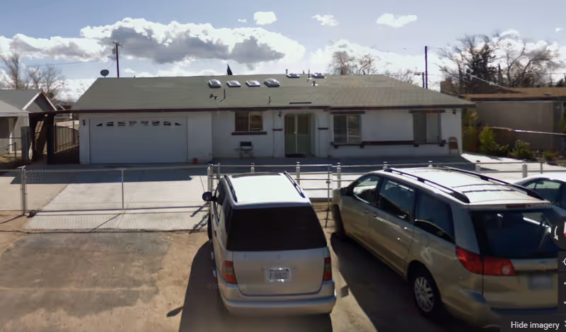 Front exterior view of a single-story residential building with a green roof, white walls, and a two-car garage. There is a chain-link fence enclosing the front yard and two parked vehicles in the driveway. The sky is partly cloudy.