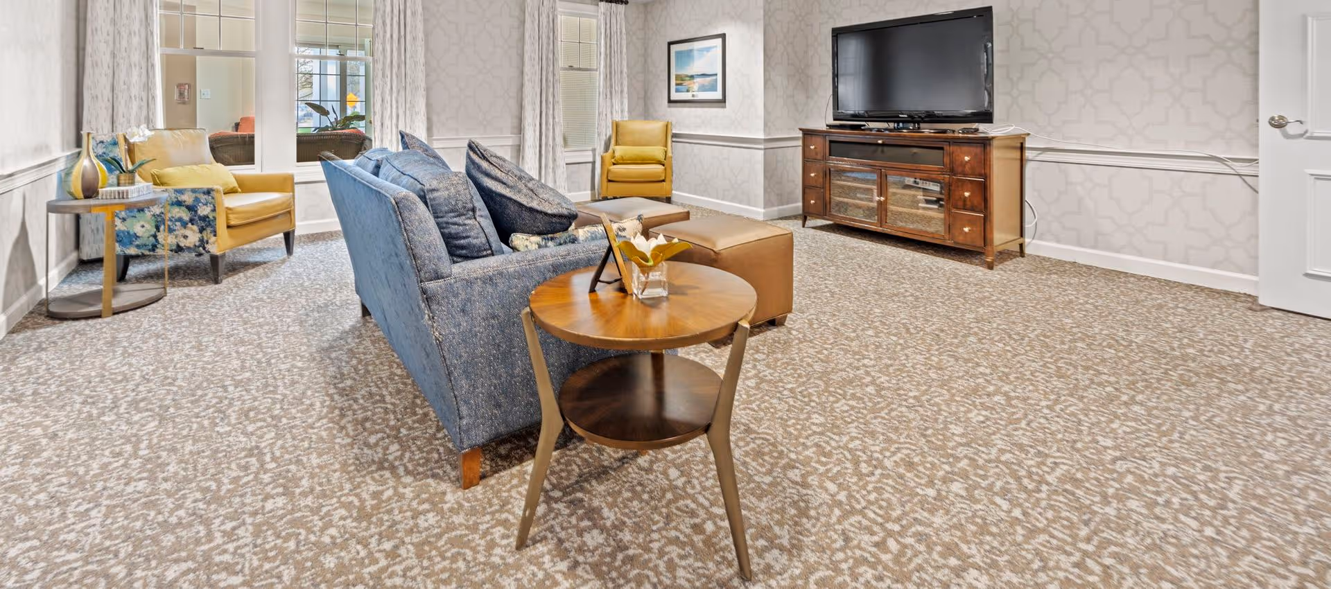 A cozy living room with a blue sofa, two yellow armchairs, a wooden side table with a small flower vase, a large flat-screen TV on a wooden TV stand, patterned carpet, and light-colored walls with a geometric pattern. There are windows with white curtains letting in natural light.