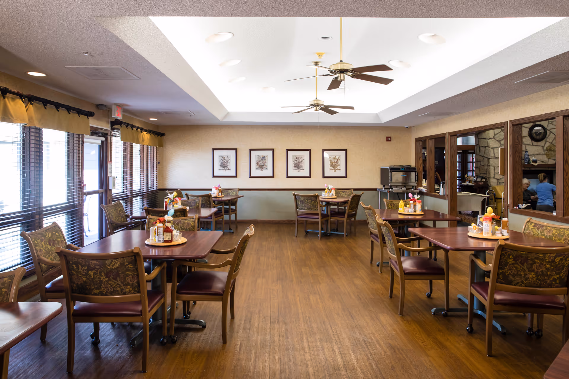 A bright dining room with several wooden tables and chairs arranged neatly. Each table has condiments and napkin holders. Large windows with blinds and valances let in natural light on the left side. The ceiling has recessed lighting and two ceiling fans. On the far wall, there are four framed botanical prints. To the right, a windowed partition reveals another room with a stone fireplace and people seated inside.