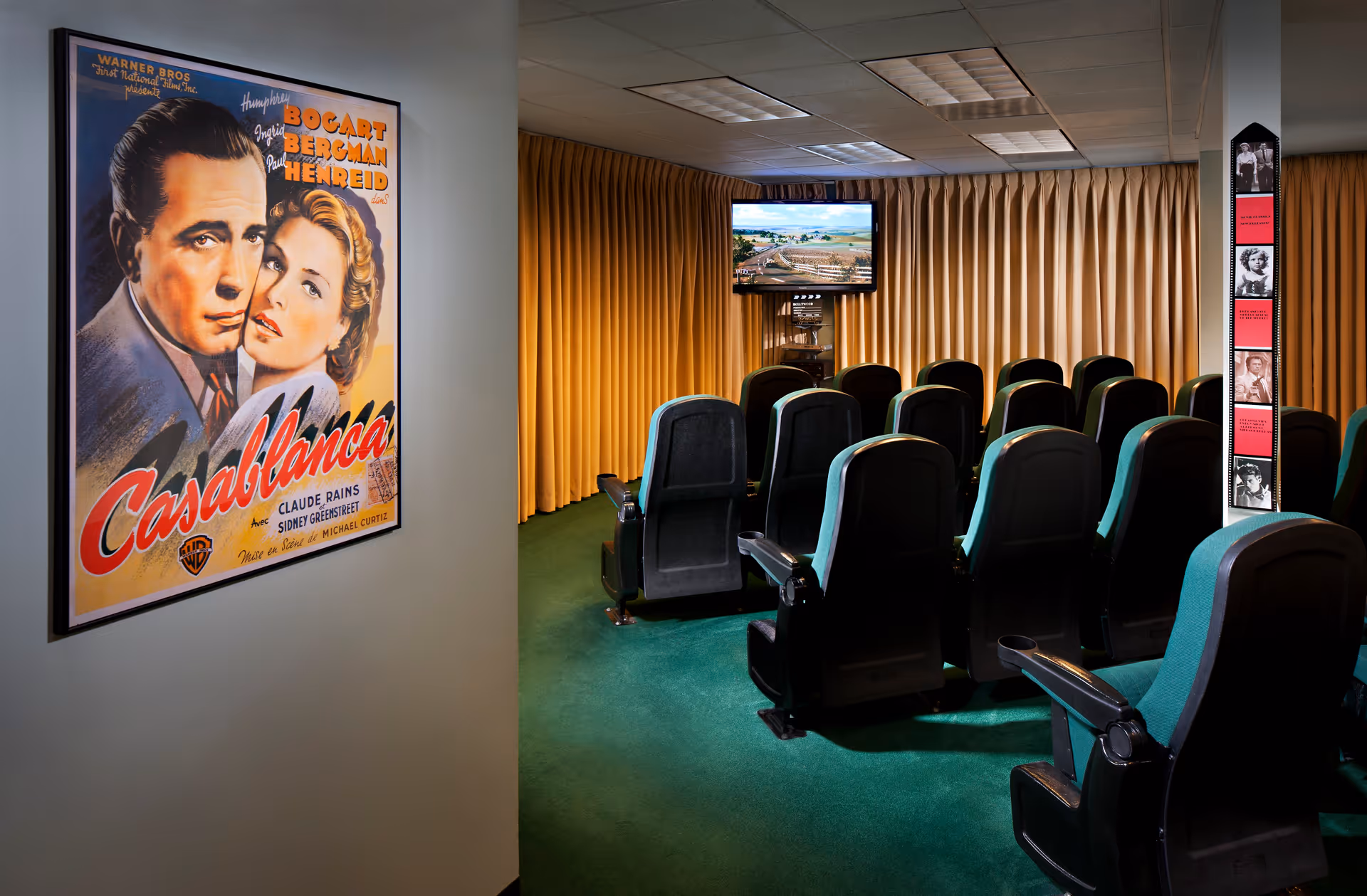 A small movie theater room with rows of green and black cushioned seats facing a wall-mounted TV screen. The room has green carpet and yellow curtains covering the windows. On the left wall, there is a framed vintage Casablanca movie poster featuring Humphrey Bogart and Ingrid Bergman.