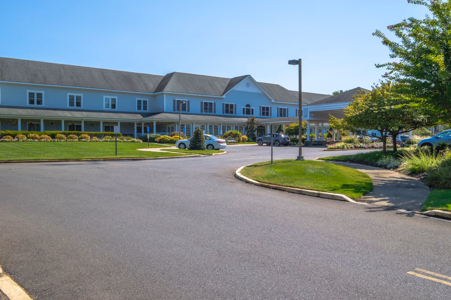Front driveway and entrance of a two-story senior living building with landscaped lawn and parked cars.
