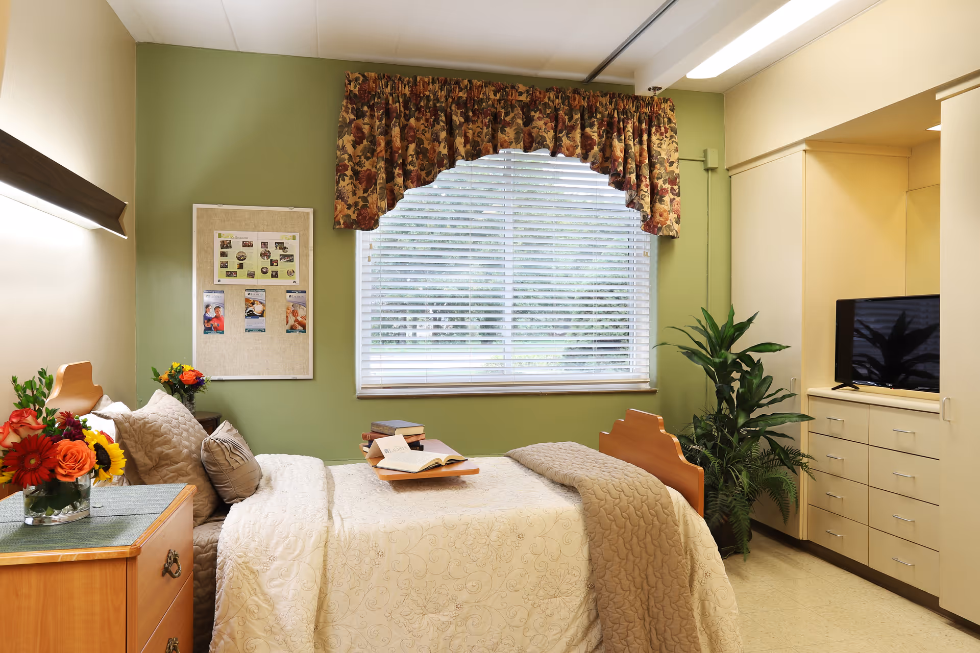 A cozy bedroom in a senior living facility with a single bed covered in a cream quilt and beige blanket. There are multiple pillows on the bed and a wooden tray with an open book and other books. A wooden nightstand with a vase of colorful flowers is beside the bed. The walls are painted green, and a large window with white blinds and floral valance lets in natural light. A bulletin board with photos and notes is on the wall next to the bed. Across from the bed is a built-in cabinet with a flat-screen TV and a green potted plant beside it.
