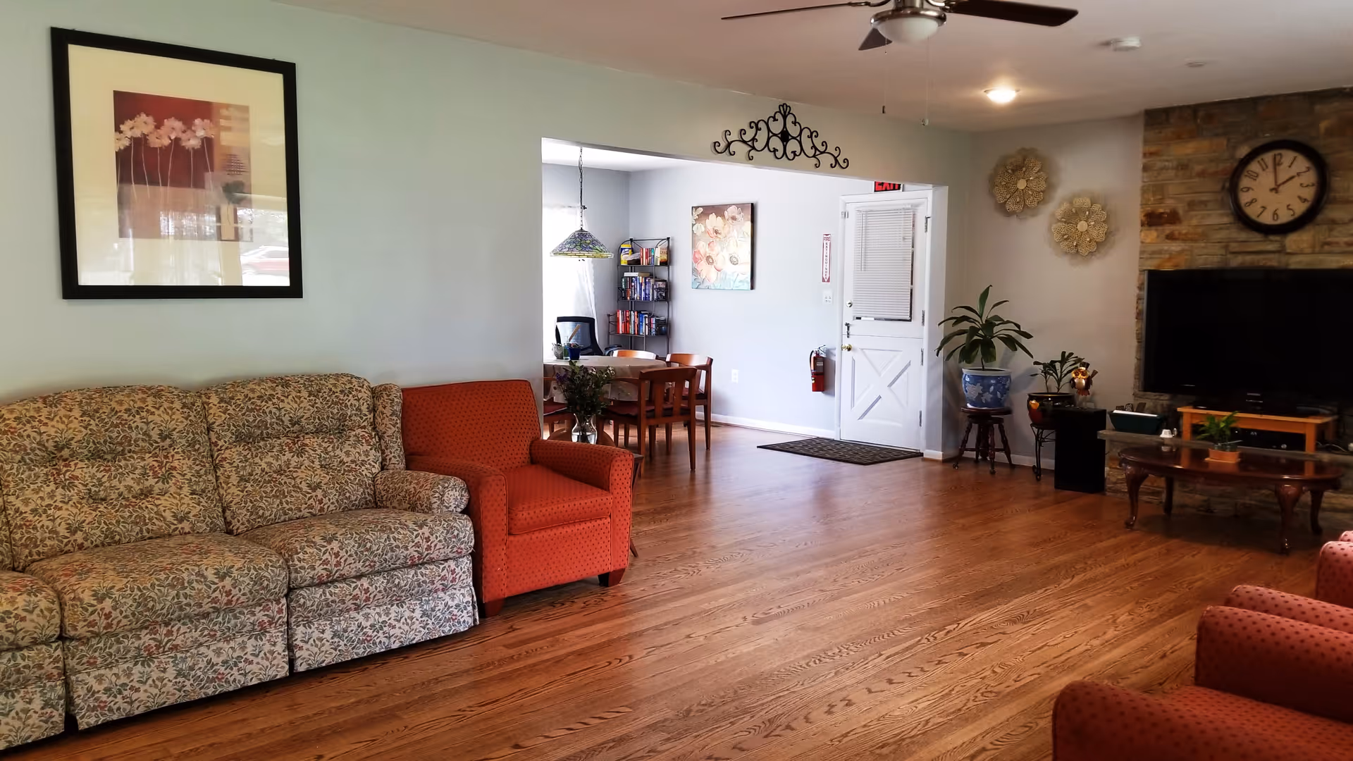 A cozy living room area with a floral patterned sofa and a red armchair on wooden flooring. The room features a ceiling fan, a wall clock above a stone fireplace, a flat-screen TV on a wooden stand, and decorative wall art. In the background, there is a dining area with a table, chairs, and a bookshelf near a white door.