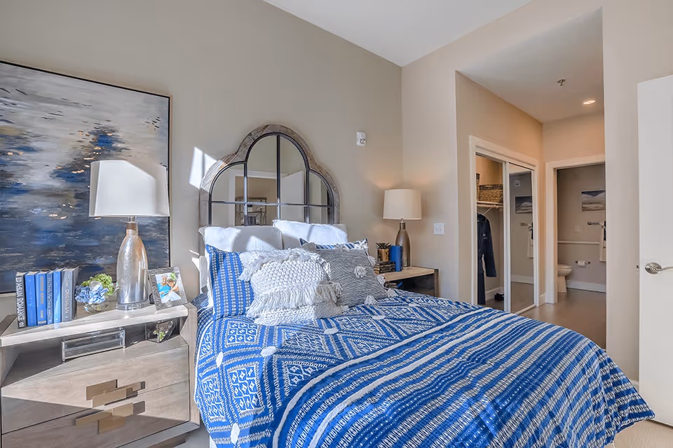 A bright bedroom featuring a bed with a blue and white patterned comforter and multiple decorative pillows. On either side of the bed are nightstands with lamps, books, and decorative items. A large abstract painting hangs on the wall to the left, and a uniquely shaped mirror is mounted above the bed's headboard. In the background, there is an open doorway leading to a walk-in closet and a bathroom.