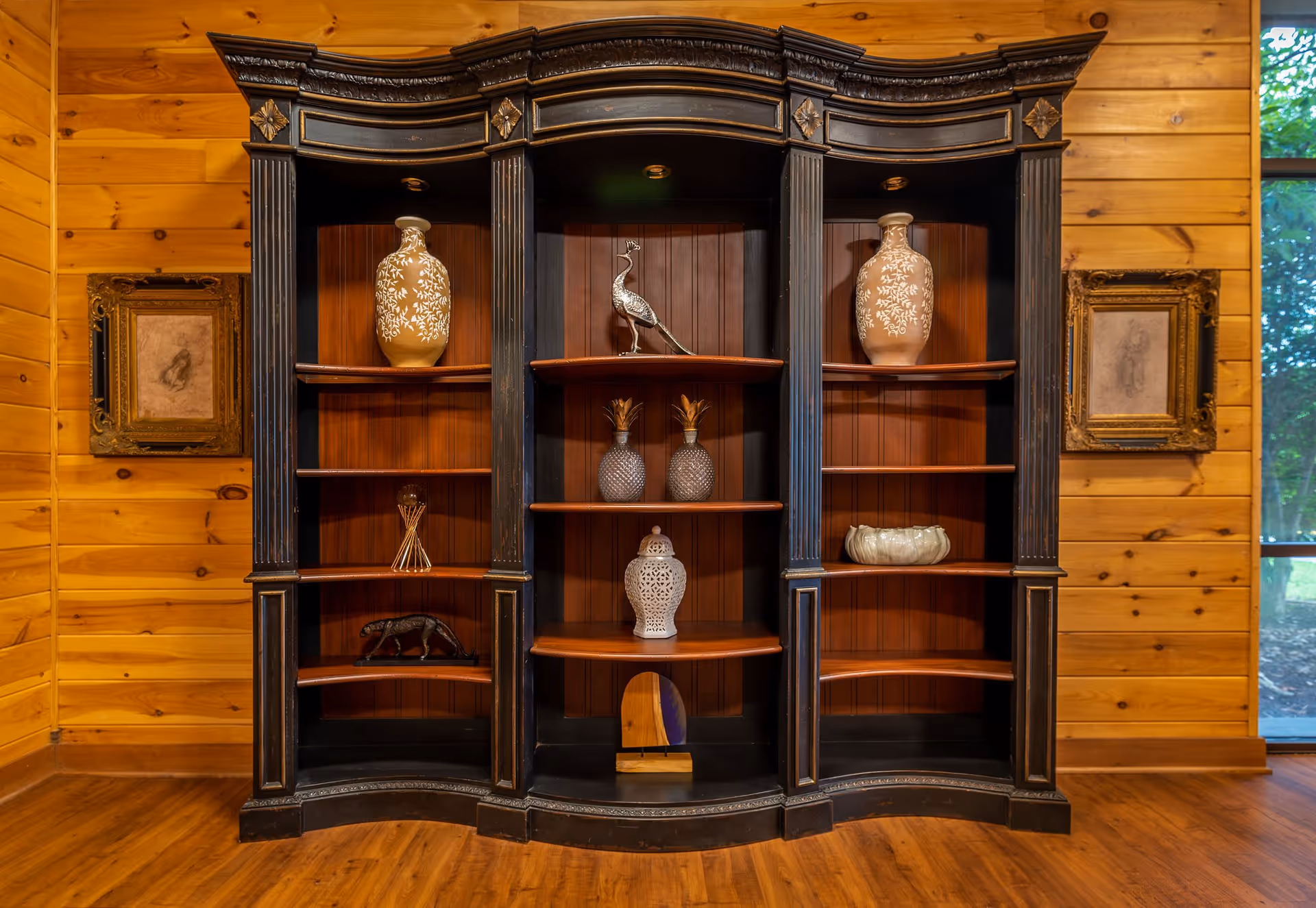 A large, ornate black wooden shelving unit with decorative vases, a silver peacock figurine, two glass pineapples, a white ceramic lantern, and other decorative items. The shelving unit is set against a wooden paneled wall with two framed sketches on either side and a window showing greenery outside.