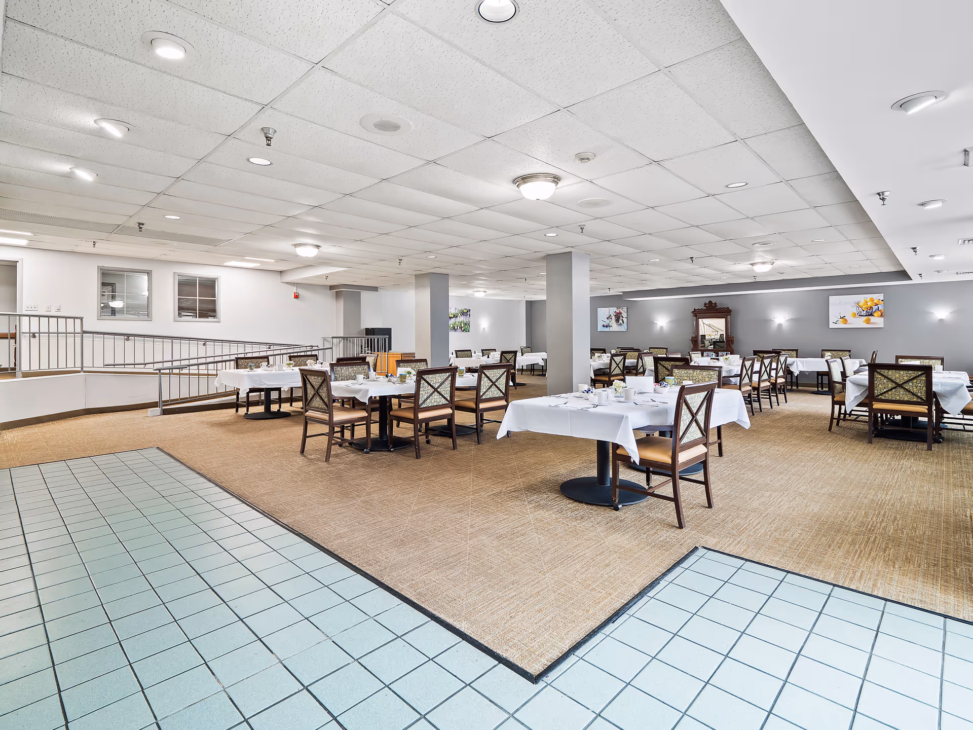 Spacious dining room with multiple tables covered in white tablecloths and set with cups and plates. The room has beige carpet flooring with a tiled area in the foreground, gray walls adorned with framed artwork, and a ceiling with recessed lighting. Wooden chairs with patterned upholstery surround the tables.