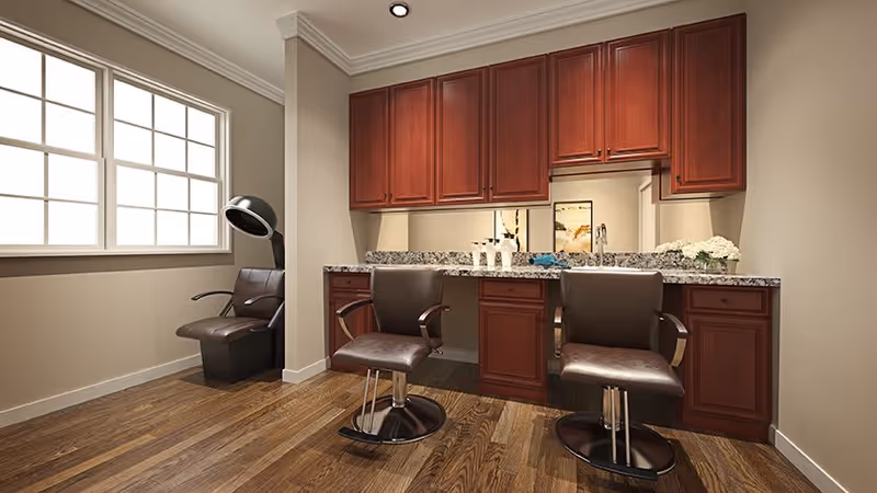 Interior view of a salon area with two brown salon chairs in front of a granite countertop and wooden cabinets. There is a large window on the left side letting in natural light, and a hair dryer chair is positioned near the window.