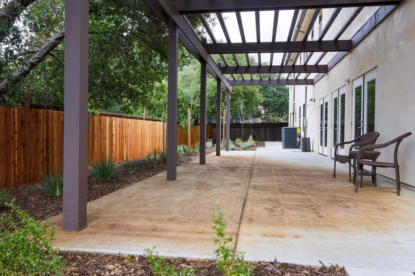 Outdoor patio area with a wooden pergola structure, concrete floor, two wicker chairs, and a wooden fence along the side. There are trees and plants surrounding the patio, and the building has multiple glass doors leading outside.