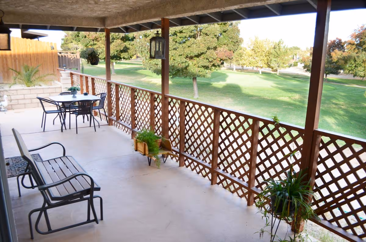 Covered patio with seating and potted plants overlooking a grassy park with trees.