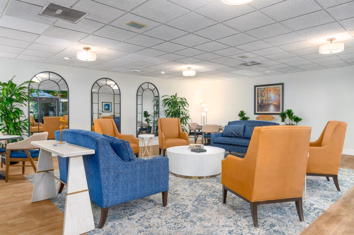 A spacious and well-lit living room area with blue and tan upholstered chairs arranged around a white circular coffee table on a patterned rug. The room features three large arched mirrors on the wall, several green plants, a framed painting, and modern ceiling lights.