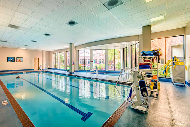 Indoor swimming pool with clear water, lane markings, and a person swimming. Large windows along one side let in natural light and show an outdoor garden area. Pool equipment and chairs are visible near the windows.