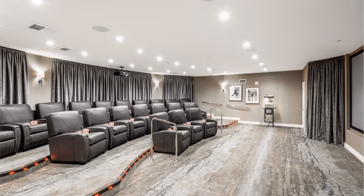 A modern senior living facility theater room with two rows of black leather recliner chairs arranged on tiered platforms. The room features a large projector screen, gray curtains covering the walls, framed artwork, and a popcorn machine in the corner. The floor has a textured carpet and the ceiling is fitted with multiple recessed lights.