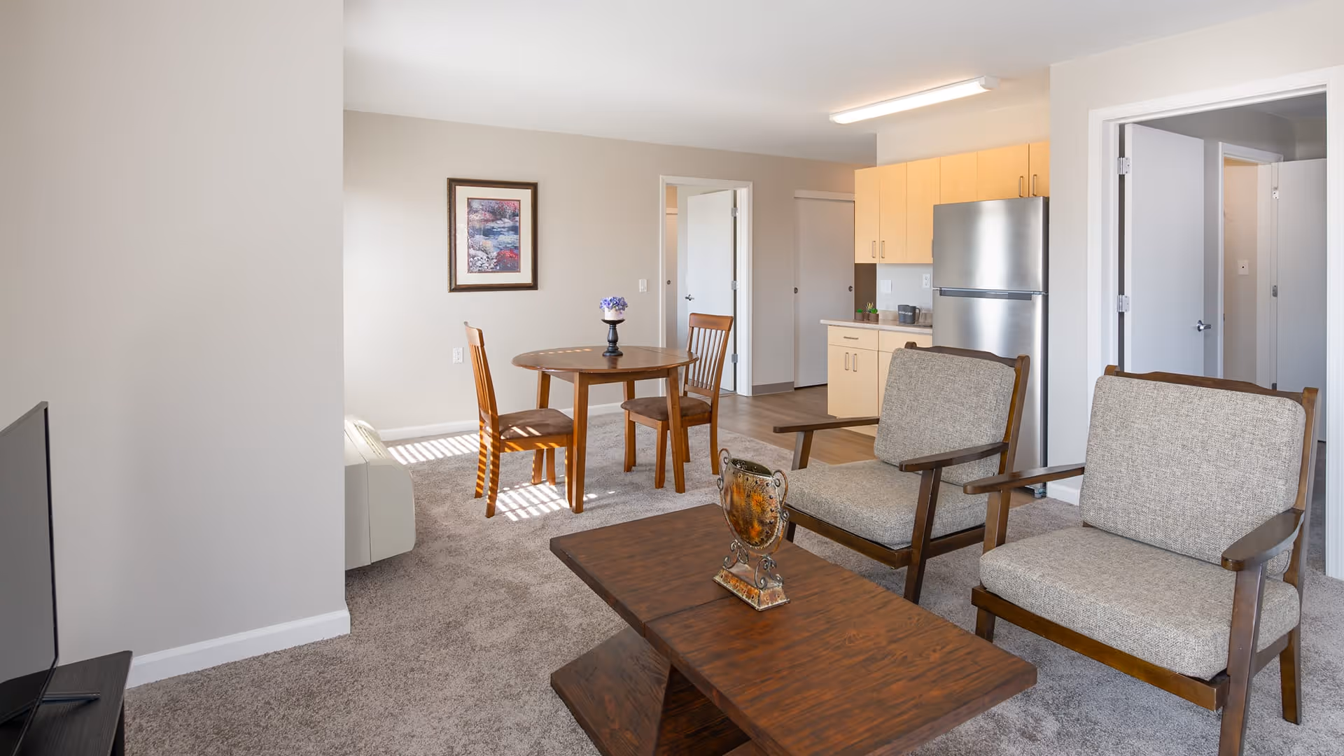 A bright and clean living area in a senior living facility featuring two cushioned wooden armchairs and a wooden coffee table with a decorative item on it. In the background, there is a small round dining table with two wooden chairs, a kitchen area with light wood cabinets and a stainless steel refrigerator, and open doorways leading to other rooms. The walls are light-colored and there is a framed picture hanging above the dining table.