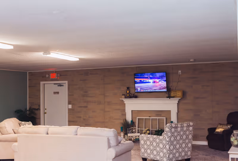A cozy living room area with beige and patterned armchairs, a white fireplace with decorative items on the mantel, a flat-screen TV mounted above the fireplace, and a door with an exit sign above it on the left side.