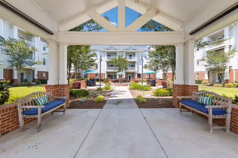 View through a covered pavilion to a landscaped courtyard with benches, umbrellas, and a three-story residential building.