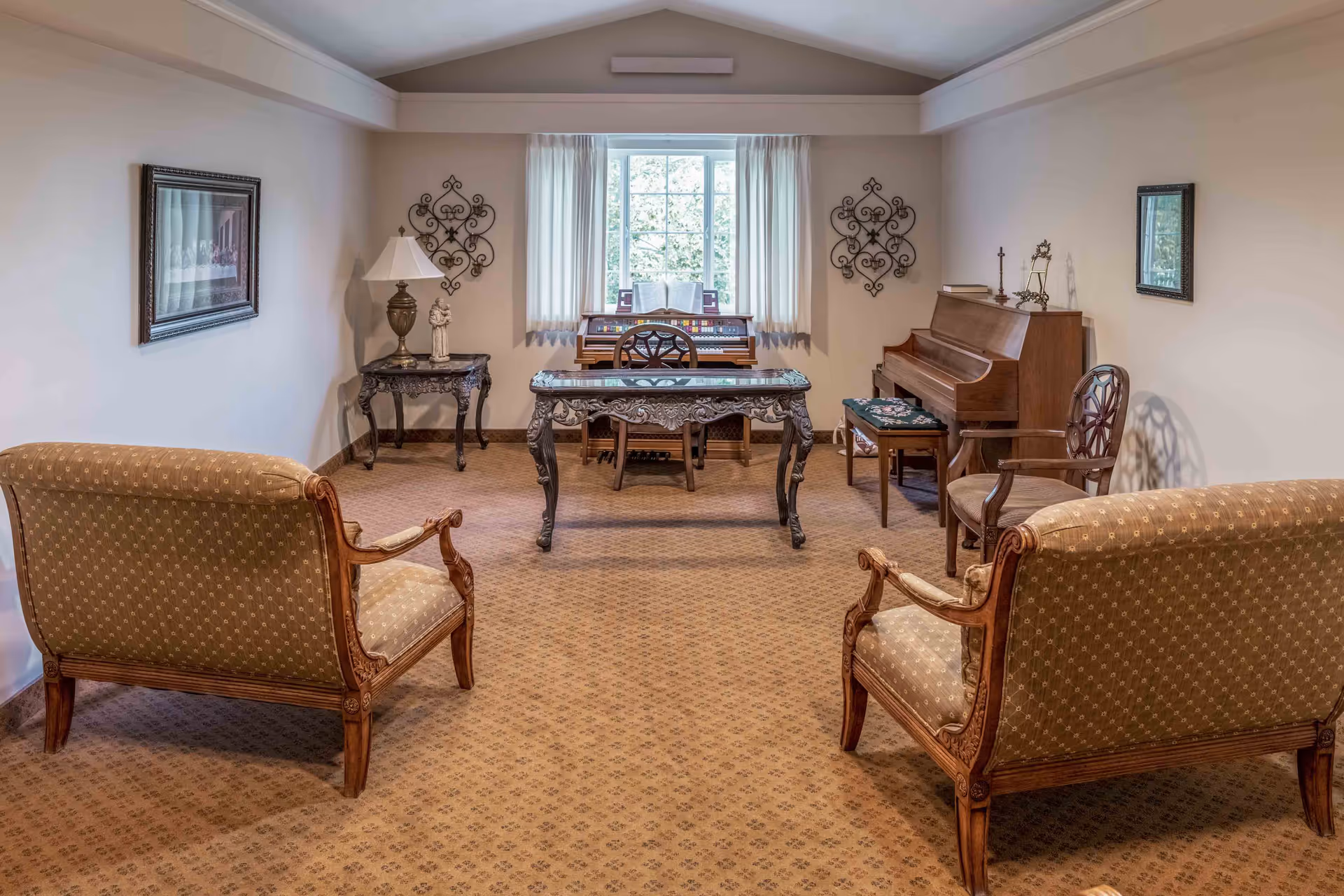 A cozy living room with two upholstered armchairs facing a decorative wooden table. In the background, there is a window with sheer curtains, an organ, and a piano with a cushioned bench. The room has beige walls adorned with framed artwork and decorative metal wall hangings, and a patterned carpet covers the floor.