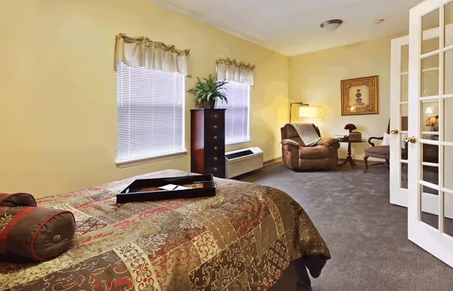 A cozy bedroom in a senior living facility featuring a bed with a patterned bedspread and decorative pillows, a wooden chest of drawers with a plant on top, two windows with blinds and valances, a comfortable recliner chair with a side table and lamp, an armchair, a framed artwork on the wall, and a glass-paneled door leading to another room.