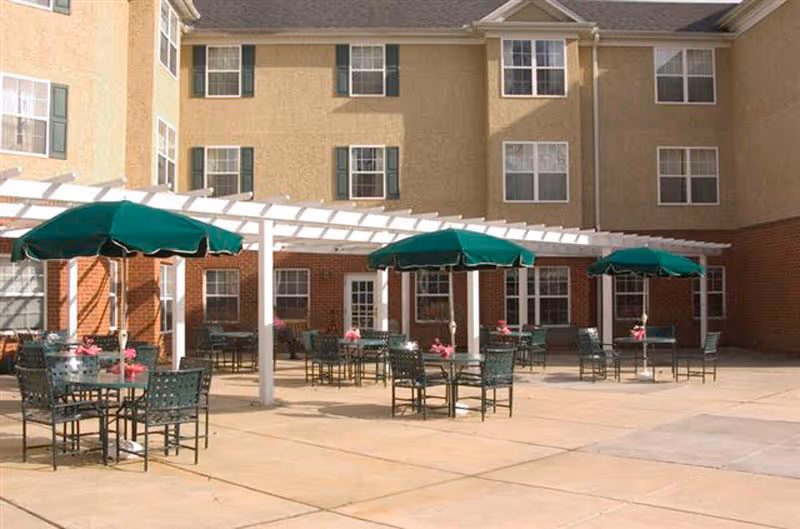 Outdoor patio area at Rockland Place with several tables and chairs under green umbrellas. The patio is paved and adjacent to a multi-story building with beige and brick exterior walls and multiple windows.