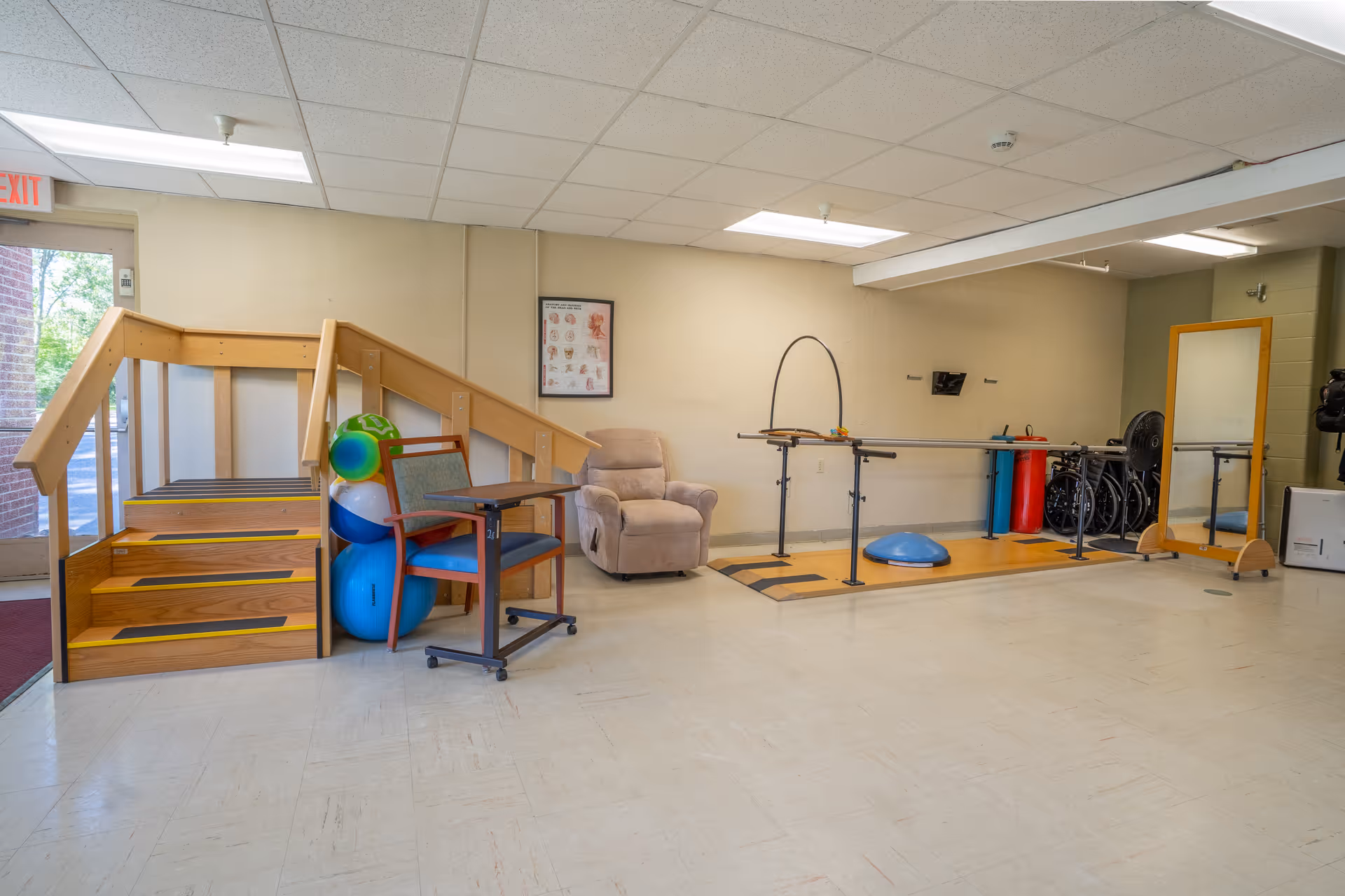 A physical therapy or rehabilitation room with wooden stairs for practice, exercise balls, a padded chair, a small table with wheels, parallel bars for walking exercises, a large mirror, and some wheelchairs in the corner. The room has a tiled floor and fluorescent ceiling lights.