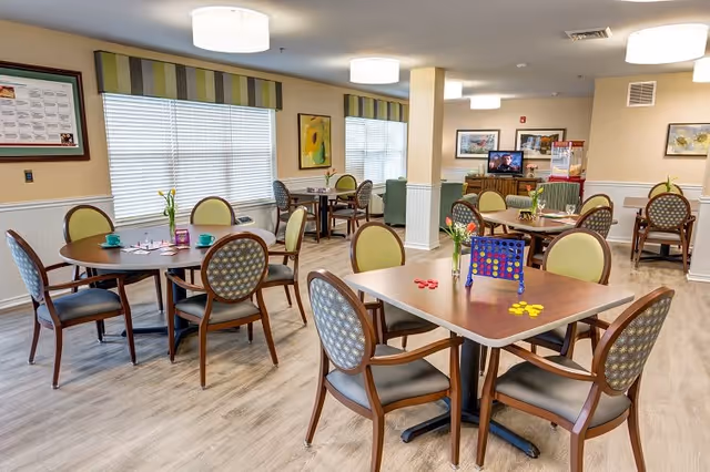 A bright and spacious dining room in an assisted living facility with several round and square wooden tables surrounded by cushioned chairs. The tables have small vases with flowers and some have board games and cups placed on them. Large windows with blinds and striped valances let in natural light. The walls are decorated with framed artwork, and the floor has light wood-style flooring.