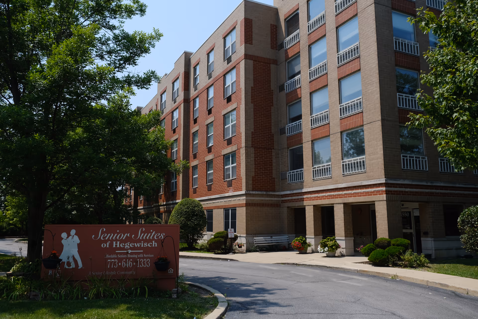 Exterior view of a multi-story senior living facility building with large windows and brick accents. There is a sign in front that reads 'Senior Suites of Hegewisch' along with contact information. The area is landscaped with trees, bushes, and flowers, and a driveway curves in front of the building entrance.
