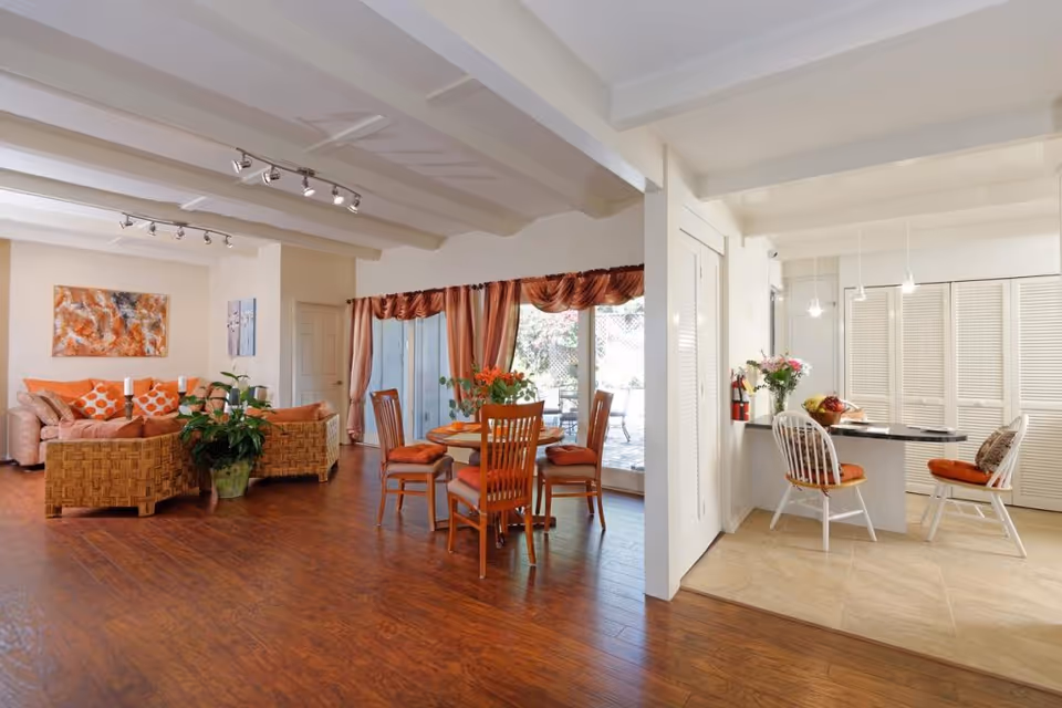 Bright open living and dining area with wicker sofas, a wooden dining table, and a small breakfast nook by the kitchen.