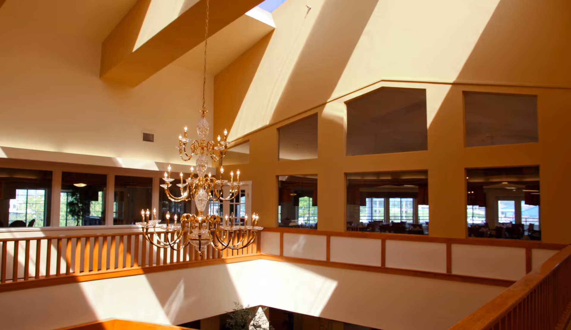 Interior view of a senior living facility with a large, ornate chandelier hanging from a high ceiling. The space is illuminated by natural light coming through skylights, casting shadows on the walls. There is a wooden railing around an open area overlooking a lower floor, with windows showing a dining area in the background.