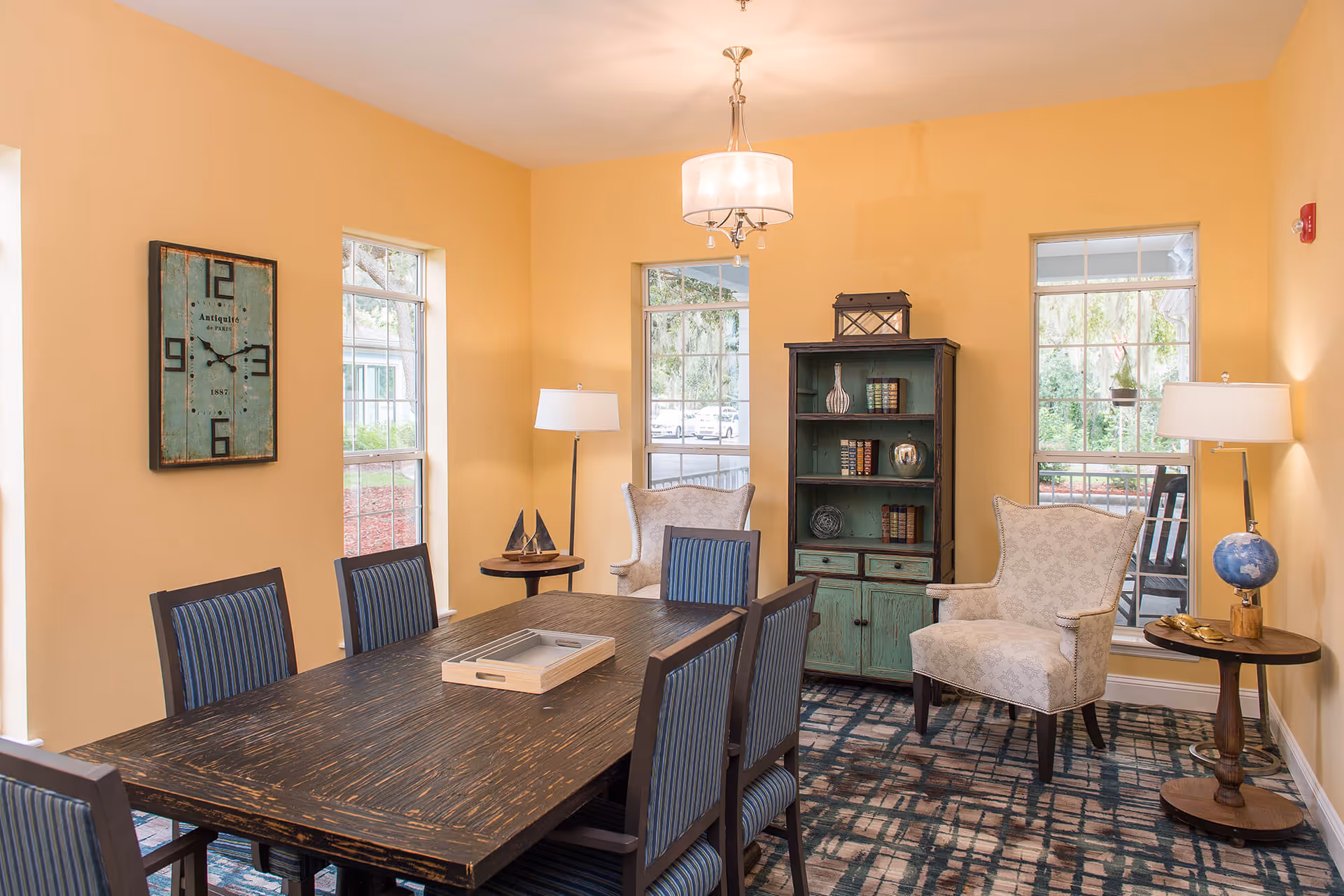 A cozy dining room with a dark wooden table surrounded by six chairs with blue striped upholstery. The walls are painted yellow, and there are three windows letting in natural light. Two beige armchairs are placed near a green distressed bookshelf filled with decorative items and books. Two floor lamps with white shades stand beside the armchairs, and a decorative clock hangs on the wall.
