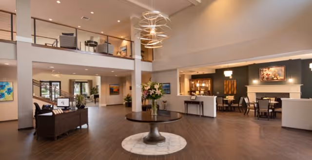 Spacious and well-lit senior living facility lobby with a round table featuring a flower arrangement in the center. The area has modern lighting fixtures, wooden flooring, and a two-story layout with a railing on the upper level. There is a seating area with a fireplace and artwork on the walls, as well as a reception desk and staircase leading to the second floor.