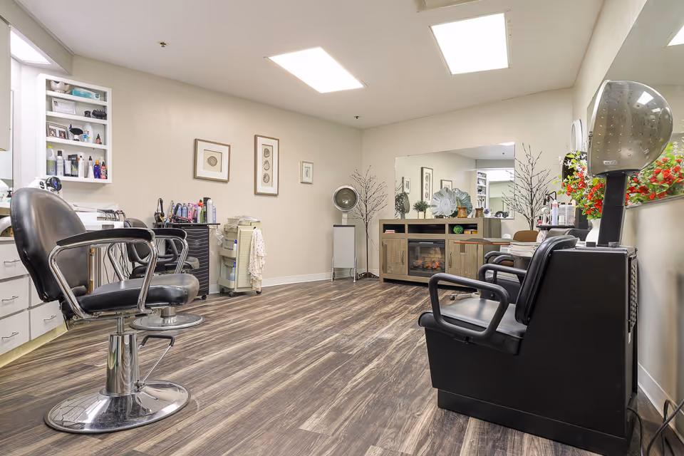Interior view of a salon area in an assisted living facility with two black salon chairs, hair care products on shelves and counters, a large mirror on the wall, framed artwork, and a wooden cabinet with decorative items and a small electric fireplace.