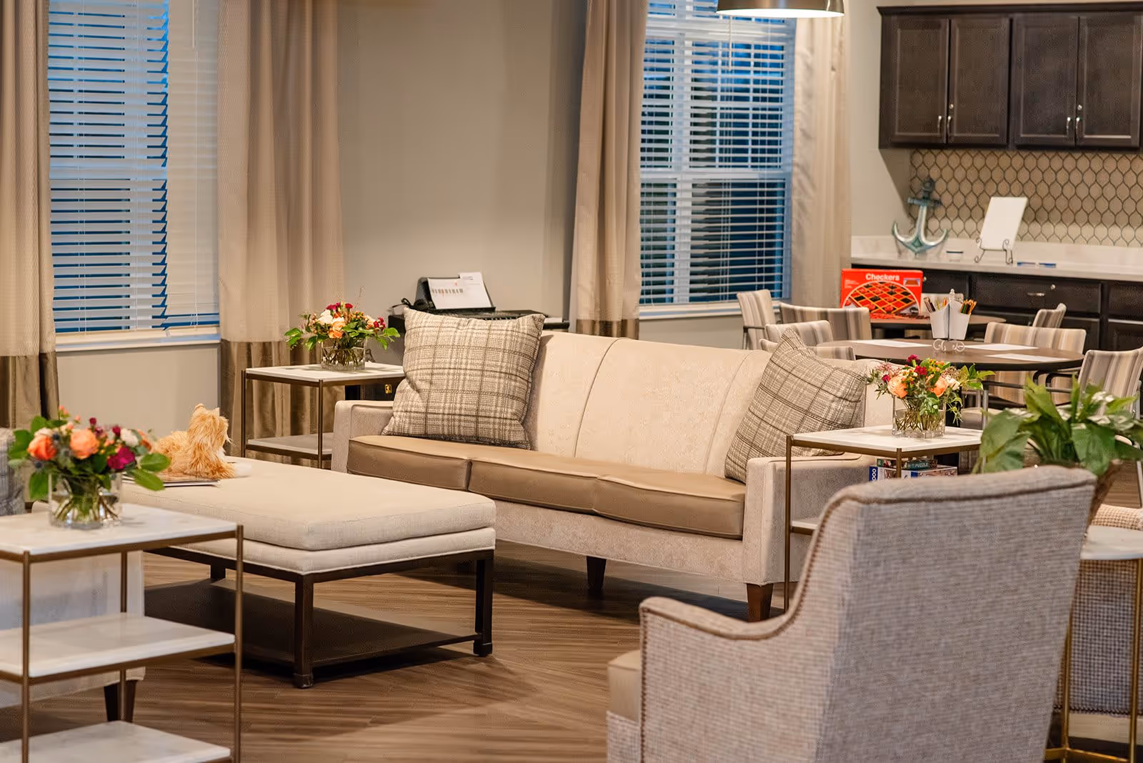 A cozy living room area with a beige sofa adorned with plaid pillows, a matching ottoman, and a comfortable armchair. There are small tables with flower arrangements and a window with blinds and beige curtains. In the background, there is a dining table with striped chairs and a kitchen area with dark cabinets and a decorative anchor.