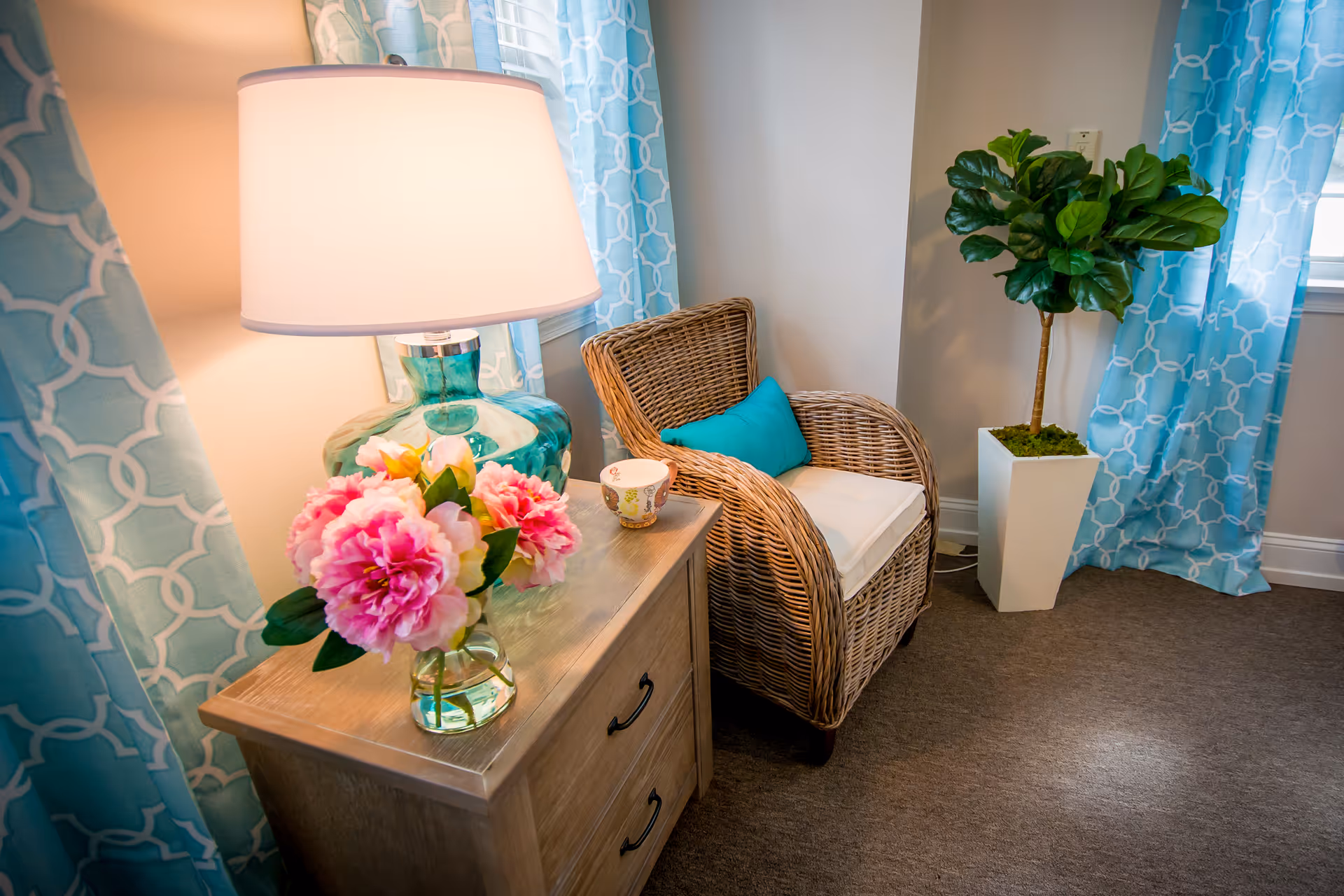Cozy corner of a room featuring a wicker armchair with a turquoise cushion, a wooden nightstand with a glass lamp and a vase of pink flowers, blue patterned curtains, and a tall potted plant in a white planter.