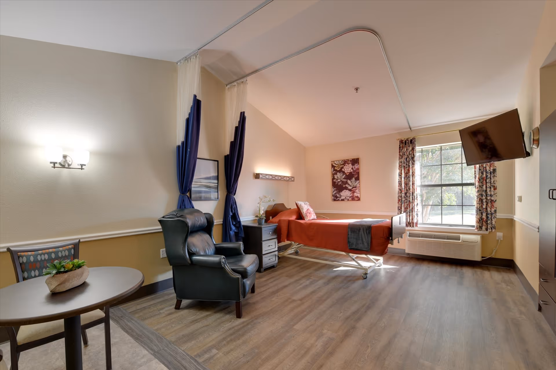 A senior living facility bedroom with a single bed covered in a red blanket and a dark throw. There is a black leather armchair next to a small table with a plant on it. The room has wood flooring, a large window with floral curtains, a wall-mounted TV, and soft lighting. A curtain track is installed on the ceiling for privacy around the bed.