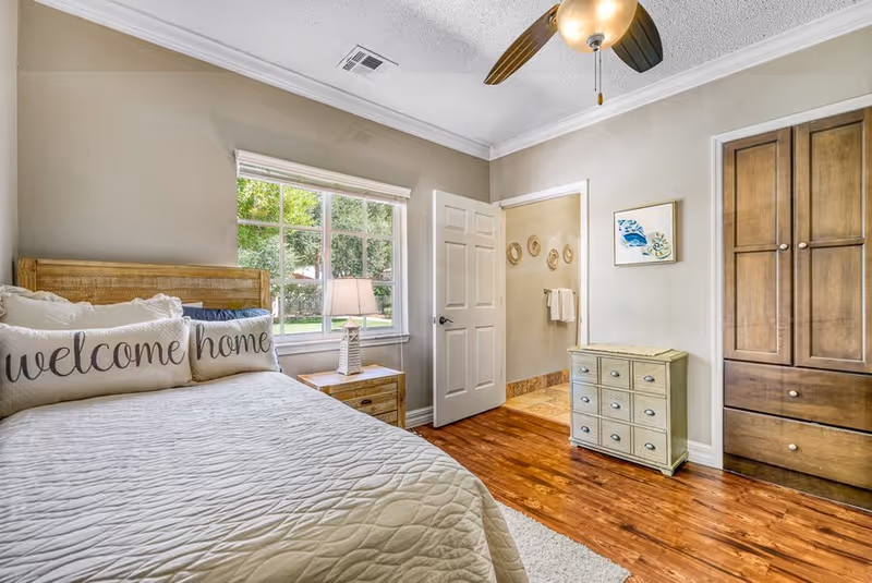 A cozy bedroom with a wooden bed frame and a quilted bedspread. Two pillows on the bed read 'welcome' and 'home'. A wooden nightstand with a lamp is next to the bed under a window showing green trees outside. The room has wooden floors, a ceiling fan with light, a small dresser, and an open door leading to a bathroom with towels hanging on the wall.