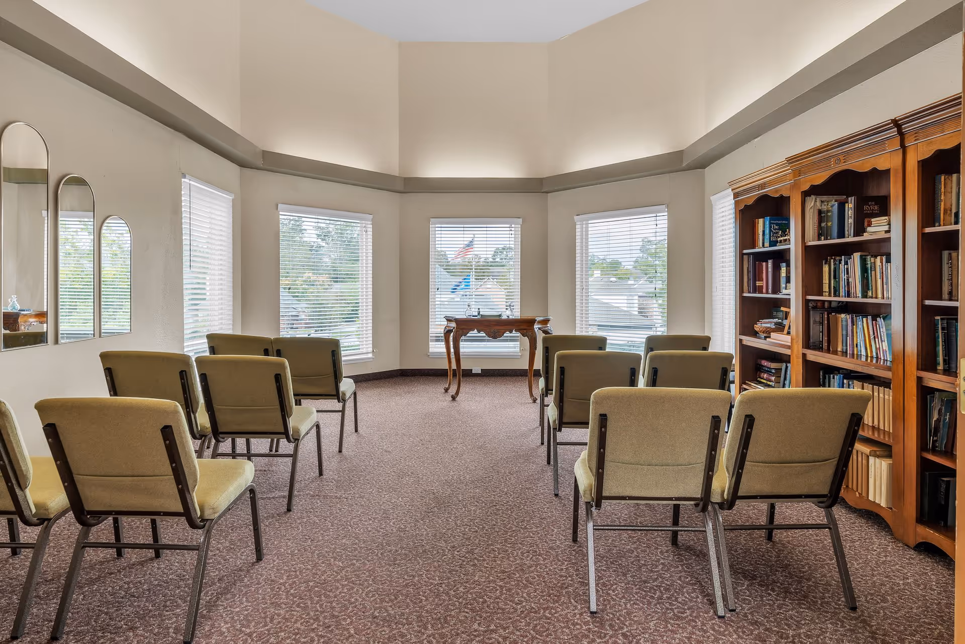 Small meeting room with rows of upholstered chairs facing a small table by bay windows and bookshelves along one wall.