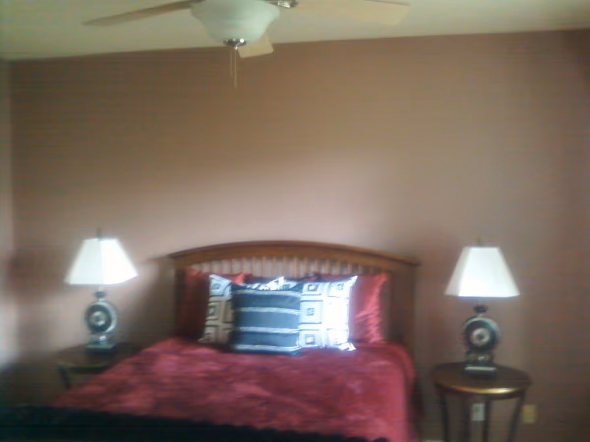 A bedroom with a wooden bed frame, red bedding, and three decorative pillows. There are two round wooden side tables, each with a lamp featuring a white lampshade. The walls are painted a muted brown color, and a ceiling fan is visible above the bed.