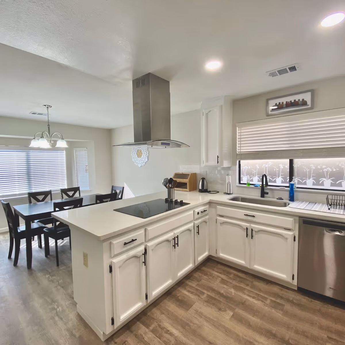 Bright open kitchen with white cabinets, a central island cooktop, stainless steel appliances and a dining table visible in the background.