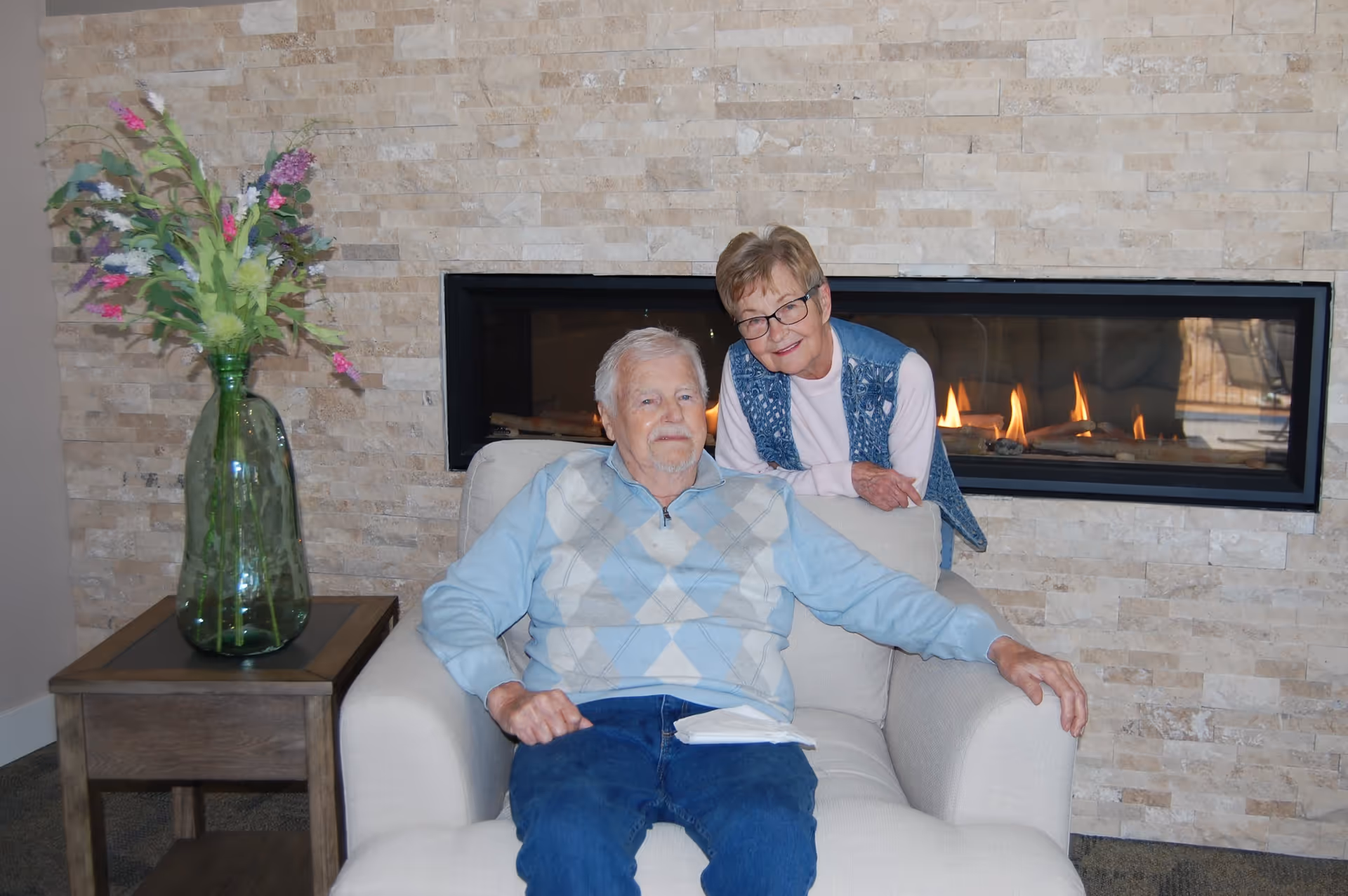 An elderly man sitting in a beige armchair with a woman standing behind him, both smiling. They are in a cozy room with a modern rectangular fireplace built into a stone wall behind them. To the left of the armchair is a wooden side table with a large green glass vase holding colorful flowers.