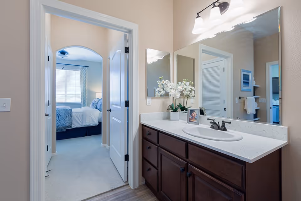 A bathroom vanity with a white countertop, dark wooden cabinets, a sink, and a large mirror above. There are decorative white flowers and a framed photo on the countertop. Through an open door, a bedroom with a bed, blue and white bedding, and a window with blinds and curtains is visible.