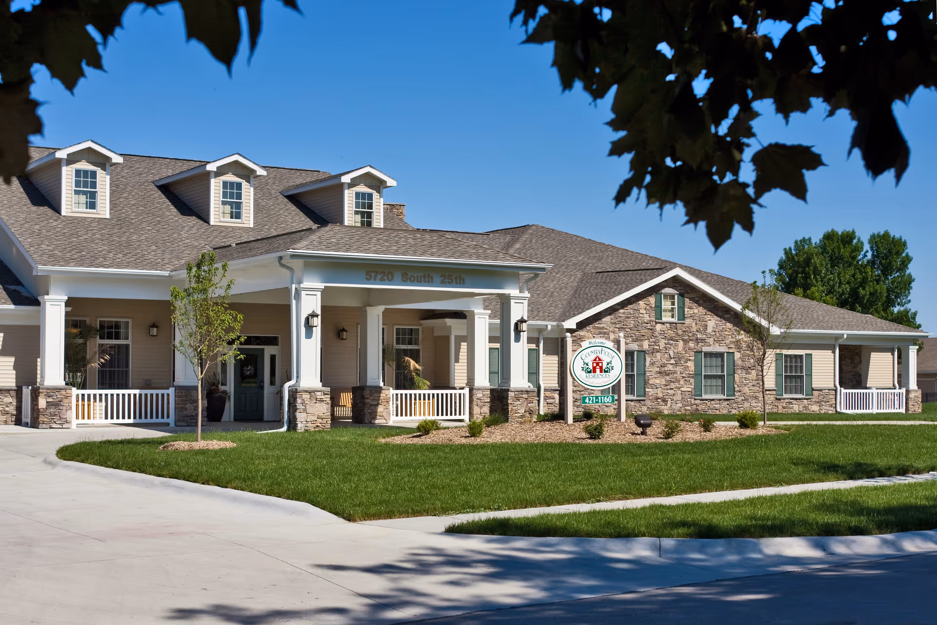 Exterior view of a single-story senior living facility building with beige siding and stone accents, a covered entrance with white columns, green doors and windows, a well-maintained lawn, and a sign displaying the facility's name and phone number under a clear blue sky.
